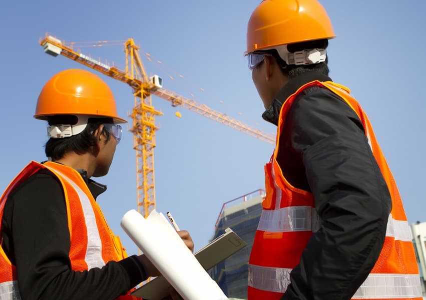Two construction workers in orange safety gear review blueprints near a large yellow crane.